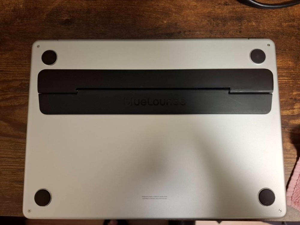 横向きの構図。木目の机の上で、シルバーのノートPC裏面に黒い Bluelounge Kickflip がヒンジ付近に中央寄せで貼付された状態。Kickflip は折りたたんだ薄型のまま