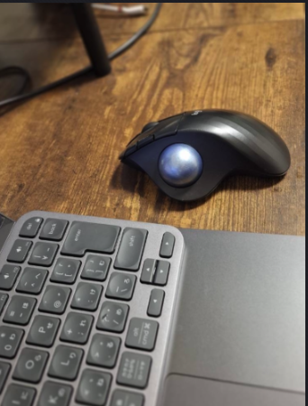 木目のデスク上に置いた、私の実機 ERGO M575。親指側の水色トラックボールと、ノートPCの日本語キーボードの一部が写っている