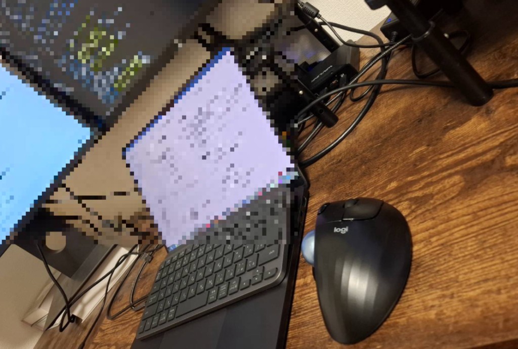 暗めの木目デスクに ERGO M575 とノートPC。奥にモニタが写る、私の実デスク俯瞰。画面はプライバシー用にぼかしている